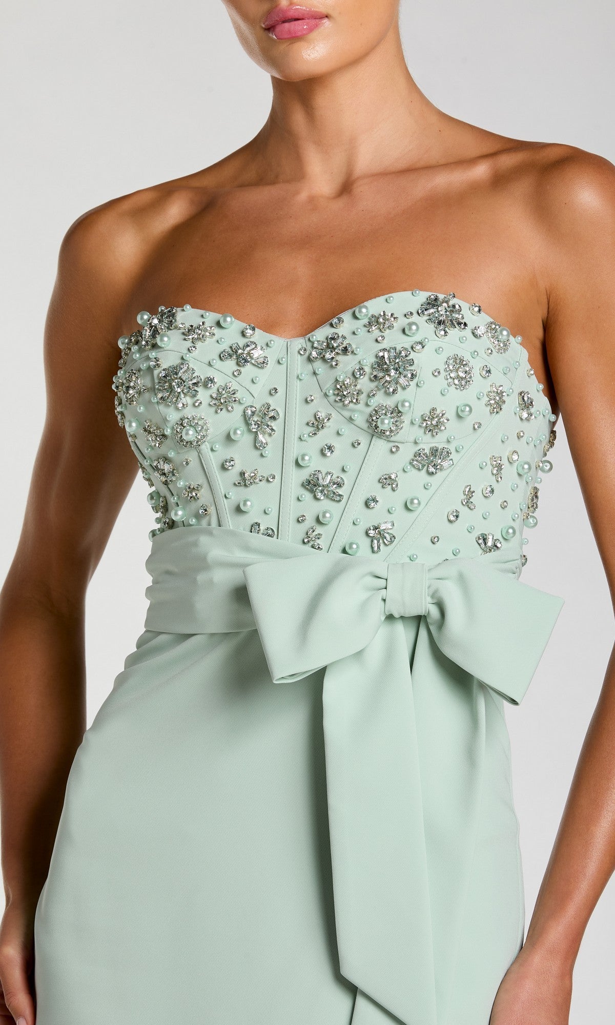 Model wears the Mac Duggal 2395 long formal dress, featuring a strapless, light green design adorned with rhinestone embellishments and a large bow at the waist; shown from shoulders to hips.
